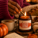 A lit amber glass jar candle labeled Natural Amie Essentials Pumpkin Vanilla Nutmeg sits on a wooden coaster, with a hand lighting it with a match. The scene is styled with a mug of hot cocoa, a small pumpkin, autumn leaves, and cozy knit fabric.