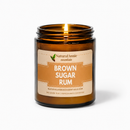 A lit scented candle in an amber glass jar with a cream and tan label reading 'Brown Sugar Rum' by Natural Annie Essentials, shown on a clean white background.